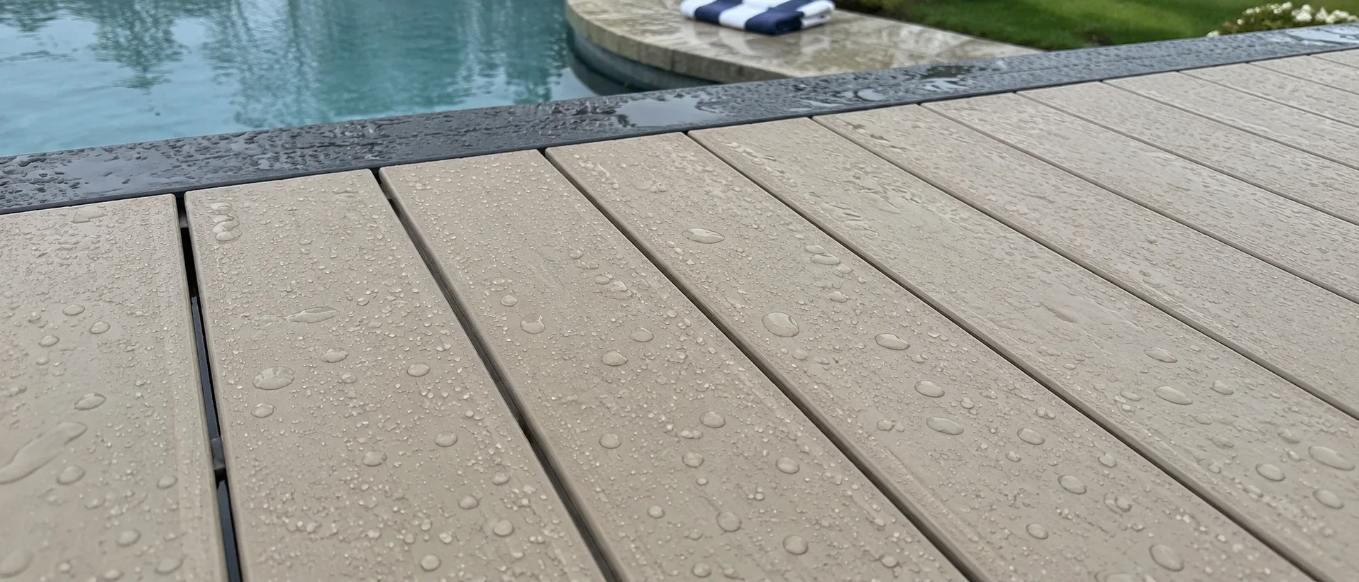 Pale capped decking boards at the edge of a swimming pool with water beading on the surface, a blue pool visible beyond the boards